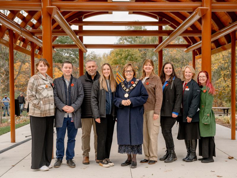 Group of people standing under the entrance of a long house structure, posing, and smiling for a picture.